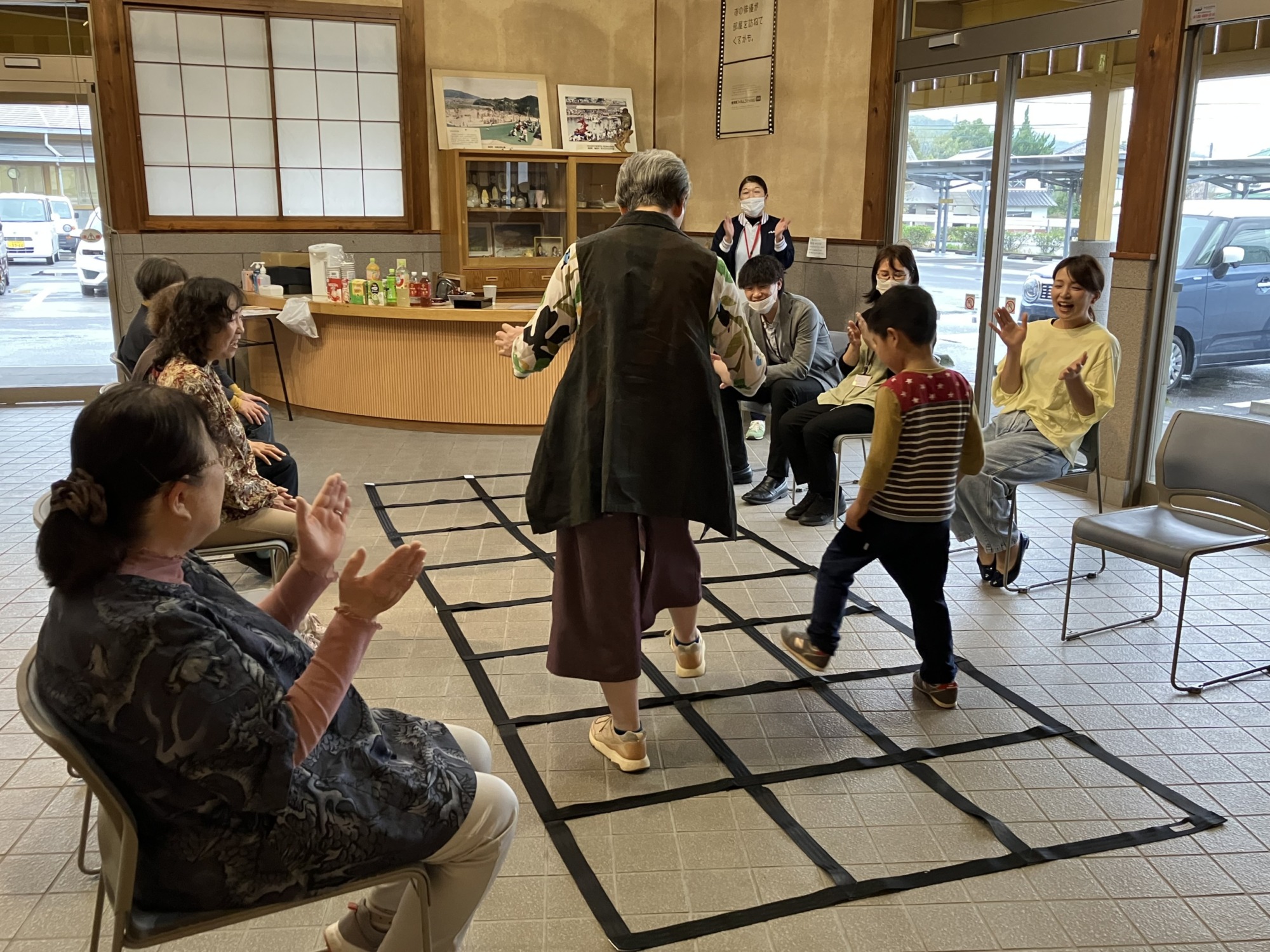 子どもと大人が地面のマス目を踏んで遊んでいる様子を椅子に座った大人たちが拍手をして見ている