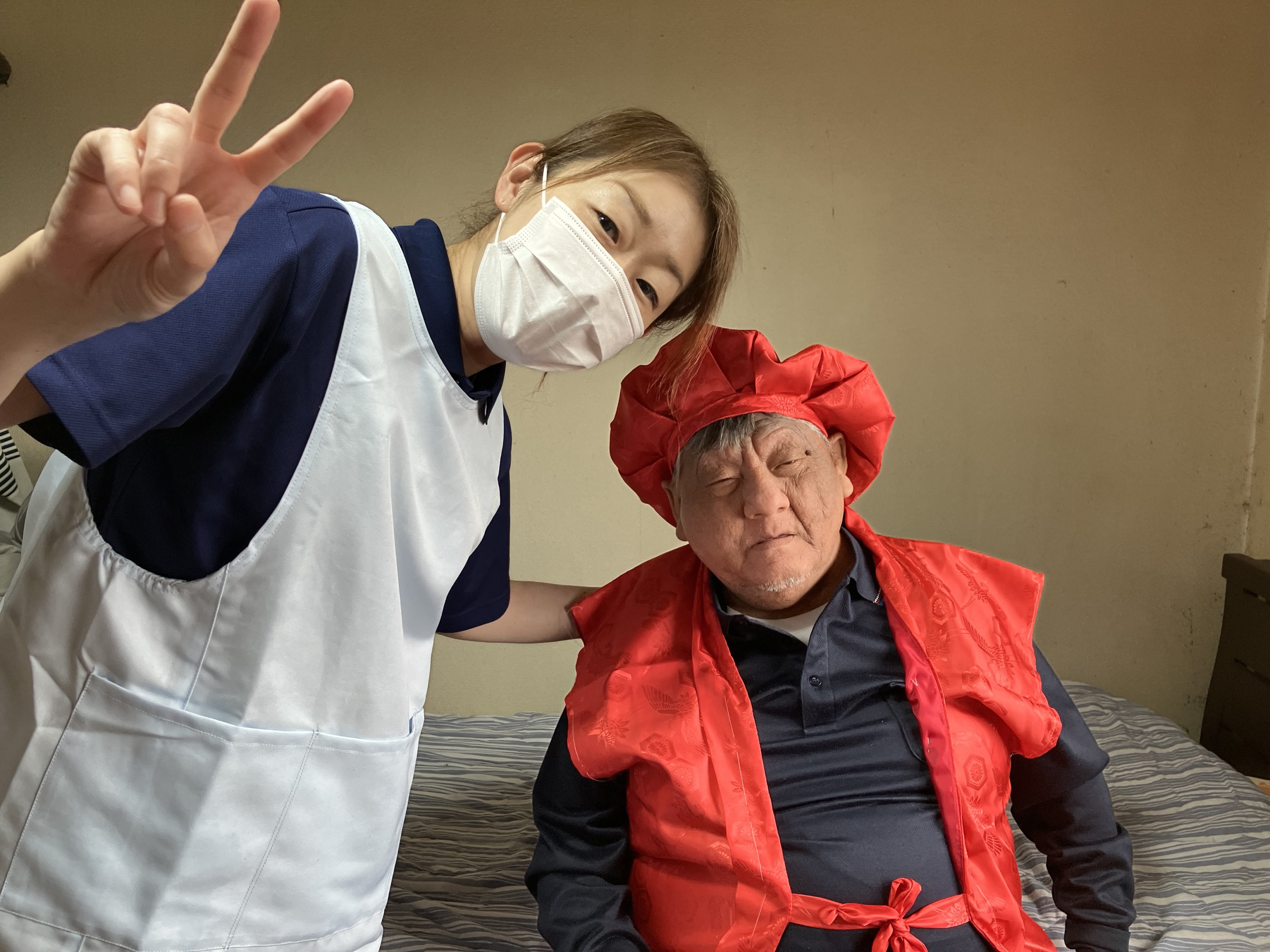 赤いちゃんちゃんこと帽子を身につけた利用者様の横でヘルパーがピースサインをしている